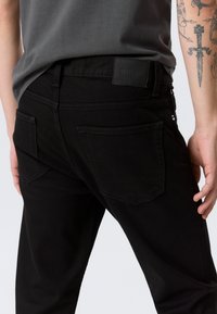Person trägt schwarze Jeans und ein dunkelgraues Hemd, zeigt die Gesäßtaschen und den Bund mit einem sichtbaren Markenpatch, tätowierter Arm.