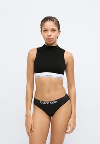 Μαύρο crop top με ψηλό λαιμό και λευκή λωρίδα με το λογότυπο Calvin Klein. Συνδυασμένο με μαύρο εσώρουχο που φέρει παρόμοιο λογότυπο. Ομαλή υφή.