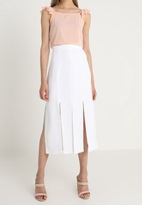 Top sans manches rose clair avec des manches volantes, associé à une longue jupe blanche avec des fentes sur le devant. Des sandales beiges neutres complètent le look.