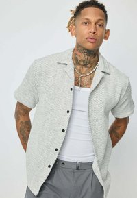 Chemise à manches courtes à boutons de couleur gris clair avec un tissage texturé, des rayures verticales et des boutons noirs. Portée par-dessus un débardeur blanc.