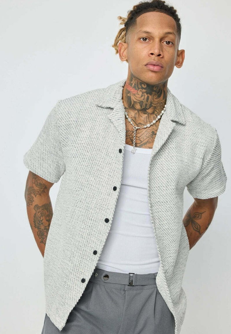 Chemise à manches courtes à boutons de couleur gris clair avec un tissage texturé, des rayures verticales et des boutons noirs. Portée par-dessus un débardeur blanc.