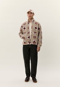 Moški, ki nosi bež vzorčast flisasti pulover, kremno kapo, črne hlače in rjave čevlje, stoji z eno roko v žepu na enobarvnem ozadju.