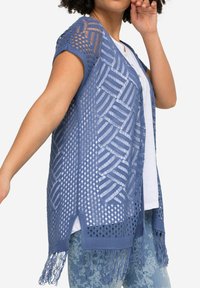 Cardigan blu leggero e a maglia aperta con motivo geometrico, maniche corte e orlo con frange. Indossato sopra una camicia bianca e jeans blu con motivo.