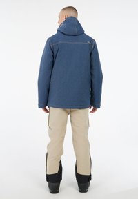 Mann mit dem Rücken zur Kamera, der eine blaue Kapuzenjacke und beige Hose mit schwarzen Stiefeln trägt, vor einem weißen Hintergrund.