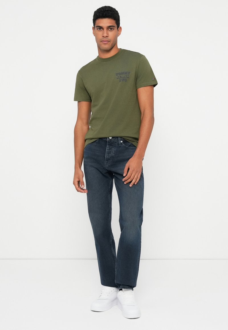 Olive green cotton t-shirt with a printed logo on the left chest, paired with dark blue jeans and white sneakers; standing pose against a white background.