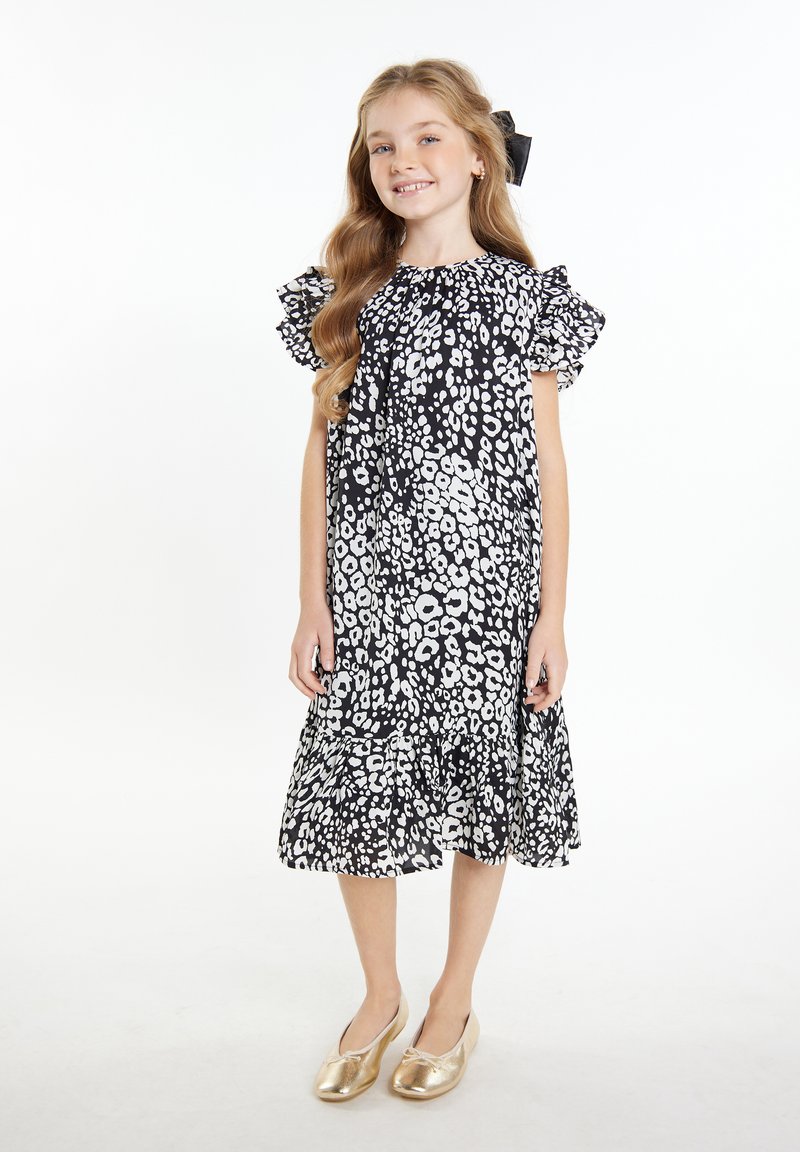 Schwarzes und weißes Blumenmuster-Kleid mit Rüschensleeves und ausgestelltem Saum. Kombiniert mit goldenen Slippern. Glatte Stoffoberfläche.