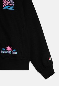 Sweatshirt noir avec une texture douce, affichant des graphismes colorés : un soleil et des vagues sur la poitrine et "sunshine mind" sur l'ourlet. Logo Champion sur la manche.
