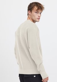 Helles, beiges Strickpullover mit strukturiertem Muster, langen Ärmeln und Rundhalsausschnitt. Der Saum ist gerippt und der Schnitt ist lässig.