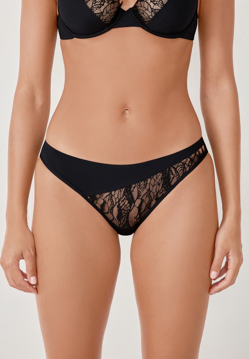 Bikini in pizzo nero con un elastico opaco e liscio. Presenta un intricato dettaglio in pizzo ai lati, evidenziando una texture trasparente e un delicato motivo.