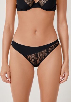 Donna che indossa un reggiseno in pizzo nero e un perizoma abbinato con dettagli in pizzo, mostrata dalla parte centrale del petto fino all'altezza delle cosce su uno sfondo neutro.