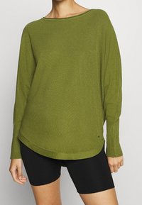 Pull en tricot vert oversize avec un décolleté bateau, tissu texturé et épaules tombantes, associé à un short de vélo noir.