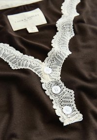Tessuto marrone con bordo in pizzo bianco e due bottoni bianchi. Presenta un design a V con dettagli in pizzo intricati lungo il colletto.