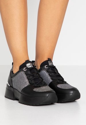 Stopy w czarnych i srebrnych błyszczących sneakersach na platformie z czarnymi sznurowadłami na białym tle.