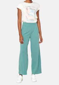 Camiseta ligera de color marfil con diseño abstracto en tonos pastel, combinada con pantalones de pierna ancha en color verde azulado y zapatillas blancas. Textura suave, ajuste casual.