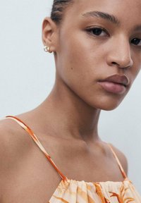 Gros plan d'une personne portant une robe légère orange à motifs avec des bretelles fines. Présente une peau lisse, des lèvres subtiles et de petites boucles d'oreilles.
