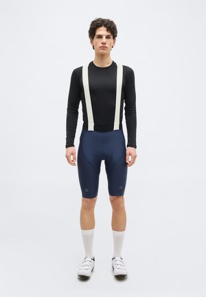 Mannlig syklist iført svart langermet topp, marineblå bib-shorts med hvite seler, hvite sokker og hvite sykkelsko, stående på hvit bakgrunn.