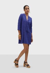 Blå tunika klänning med trekvartsärmar, med en strukturerad diamantmönster och tofsdetaljer, tillsammans med svarta plattformssandaletter.