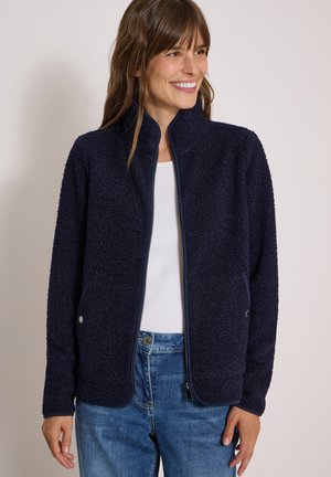 Femme souriante portant une veste polaire texturée bleu marine zippée sur une chemise blanche et un jean bleu sur un fond uni.