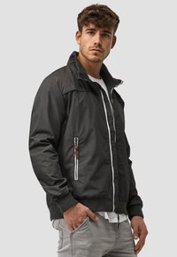 Black bomber jacket with zippered pockets and brown leather accents, featuring a stand-up collar and ribbed cuffs, made from a smooth fabric.