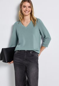 V-Ausschnitt, Top mit drei Viertelärmeln aus weichem Teal-Stoff, kombiniert mit hochtaillierten schwarzen Jeans. Hält eine schwarze Clutch mit mattem Finish.