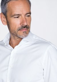 Camicia bianca con colletto a punta, tessuto liscio e vestibilità su misura. Cuciture ordinate e una leggera lucentezza sono visibili.
