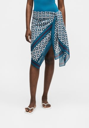 Kobieta ubrana w niebieską spódnicę kopertową do kolan z wzorem, na kostium kąpielowy w kolorze niebieskim i białe klapki, stojąca na białym tle.