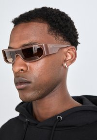 Zonnebril met een rechthoekig frame in doorzichtig bruin, getinte lenzen en subtiele branding aan de zijkant. Gecombineerd met een zwarte hoodie.