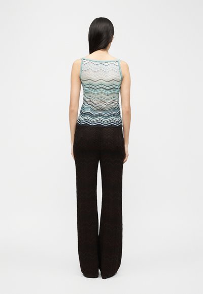 Γυναίκα με μακριά σκούρα μαλλιά, στέκεται φοράει μια πολύχρωμη μπλούζα χωρίς μανίκια με σχέδιο ζιγκ ζαγκ και μαύρο παντελόνι με φαρδιά πόδια, γυρισμένη προς τα πίσω σε λευκό φόντο.