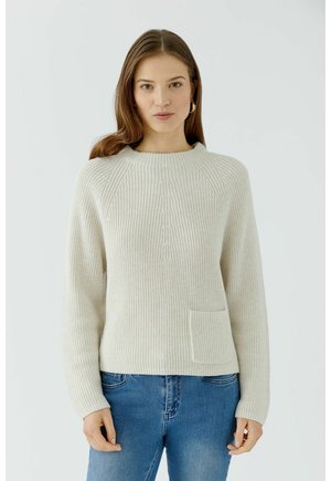 Vrouw met lang bruin haar die een crèmekleurige gebreide trui met een voorzak en een blauwe spijkerbroek draagt, staand tegen een effen lichte achtergrond.
