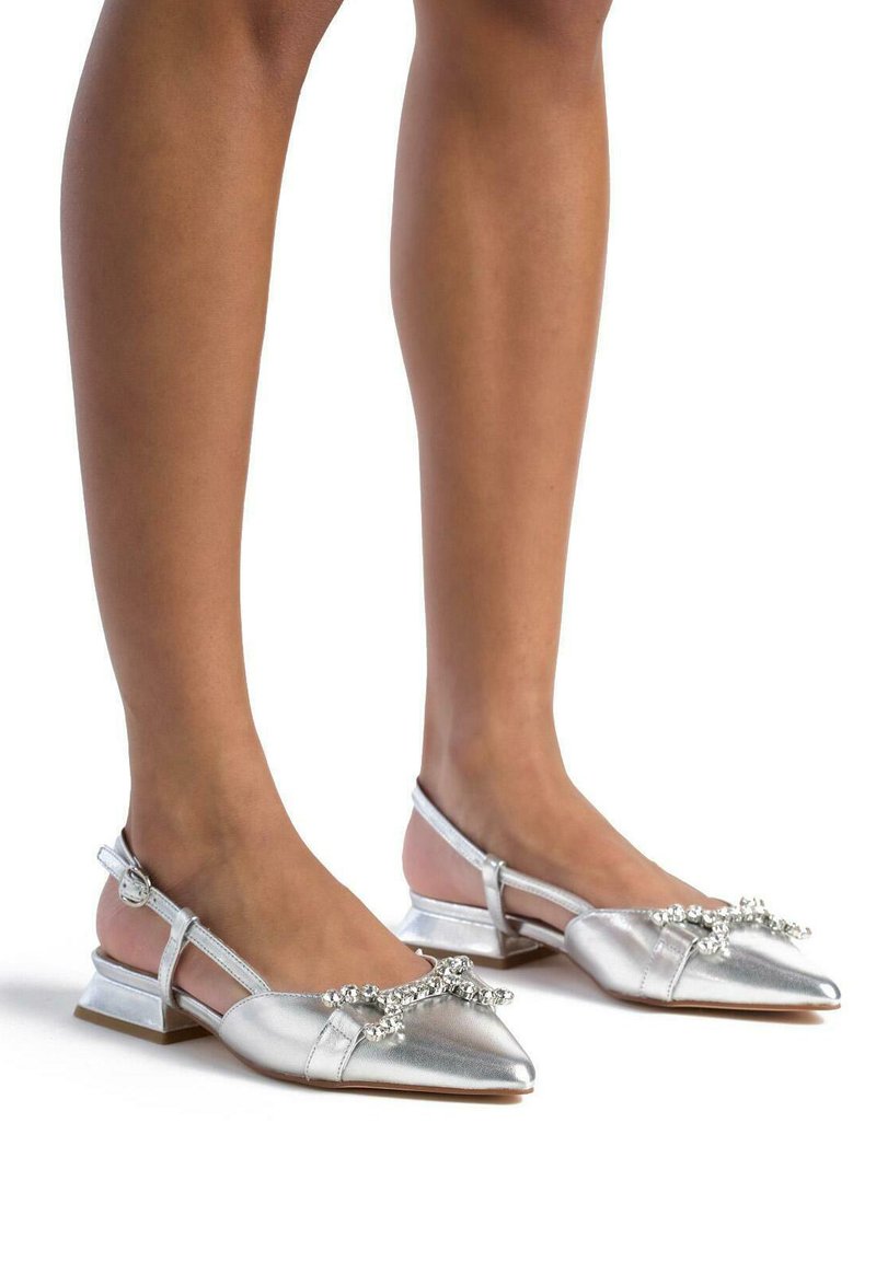 Zapatos slingback metálicos plateados con punta afilada, adornados con un lazo decorativo y piedras, y un tacón bajo y angular.