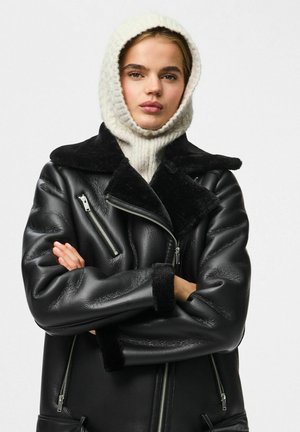 Chaqueta de cuero negra con cuello de borrego, bolsillos con cremallera y mangas anchas, usada sobre un suéter de punto ligero y ajustado con capucha.