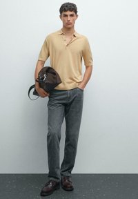 Polo beige a maniche corte, abbinato a jeans grigi e scarpe marroni. Una borsa marrone è tenuta con una mano. Sfondo semplice.