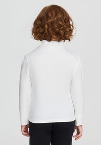 Dziecko z kręconymi czerwonymi włosami stoi tyłem, ubrane w białą bluzkę z długim rękawem i czarne spodnie na jednolitym tle.