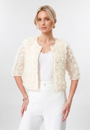 Donna con capelli biondi indossa una giacca corta color crema con trama a rose, maniche a tre quarti, pantaloni bianchi, orecchini d’oro e un braccialetto.