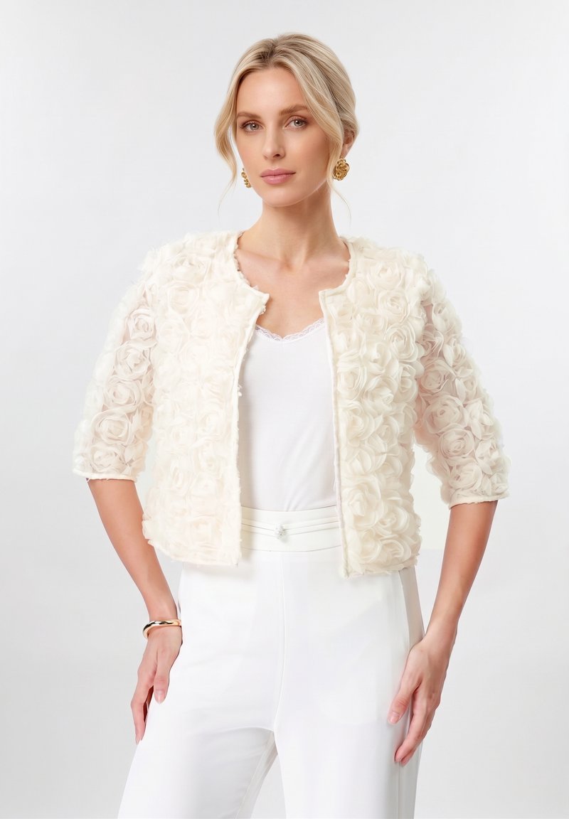 Femme aux cheveux blonds porte une veste courte texturée motif rose crème à manches trois-quarts, pantalon blanc, boucles d’oreilles dorées et un bracelet.