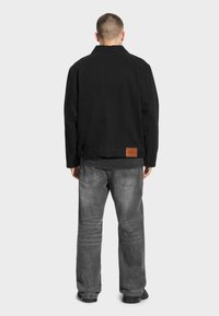 Czarna bawełniana kurtka z kołnierzykiem, w połączeniu z szarymi dżinsami. Z tyłu znajduje się brązowa skórzana metka z tłoczonym napisem "PROSTO".