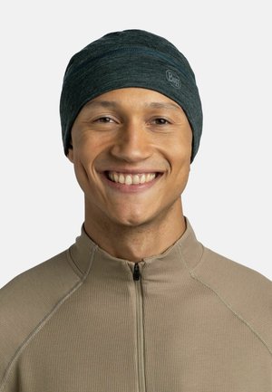 Dark teal beanie made of soft, stretchy material, featuring a subtle logo and a smooth texture, worn by a smiling person in a tan top.