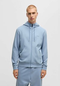 Ljusblå zip-up hoodie i mjukt tyg, med dragsko huva, sidofickor och en tonalt logotyp på bröstet.