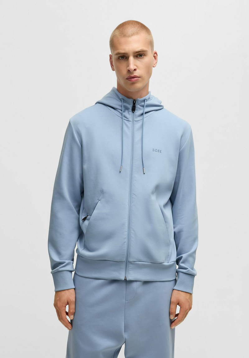 Ljusblå zip-up hoodie i mjukt tyg, med dragsko huva, sidofickor och en tonalt logotyp på bröstet.