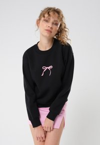 Svart genser med rosa sløyfe broderi i sentrum; har ribbestrikket rund hals og mansjetter. Brukt sammen med et rosa miniskjørt.