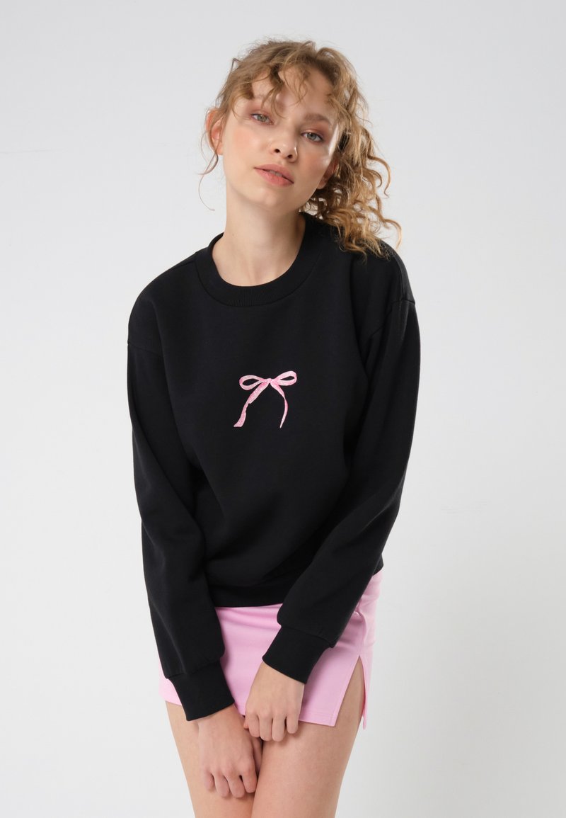Svart genser med rosa sløyfe broderi i sentrum; har ribbestrikket rund hals og mansjetter. Brukt sammen med et rosa miniskjørt.
