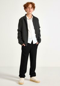 Grå zip-up jakke over en hvid skjorte med knapper, sorte bukser og beige sneakers. Jakken har en afslappet pasform og ribbede manchetter.