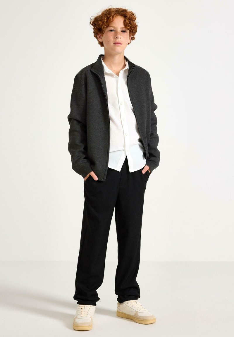 Grå zip-up jakke over en hvid skjorte med knapper, sorte bukser og beige sneakers. Jakken har en afslappet pasform og ribbede manchetter.