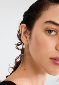 Srebrne viseče uhane krasijo dizajn v obliki solze z prozornim kamnom. Model ima valovite temne lase in posnetek v bližini ušesa.