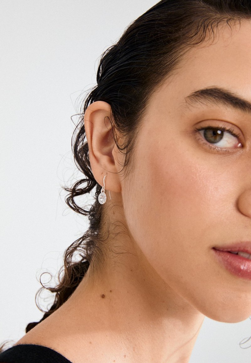 Srebrne viseče uhane krasijo dizajn v obliki solze z prozornim kamnom. Model ima valovite temne lase in posnetek v bližini ušesa.