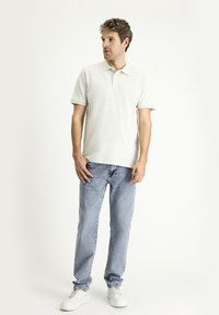 Polo shirt grigio chiaro con colletto e maniche corte, abbinato a jeans blu chiaro e sneakers bianche. Su uno sfondo uniforme.