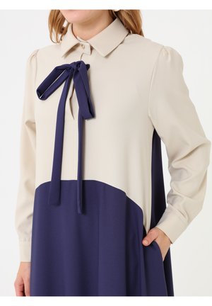 Donna che indossa un vestito beige e blu navy a maniche lunghe con colletto, fiocco blu navy all’altezza dello scollo e mano nella tasca del vestito sul lato destro.