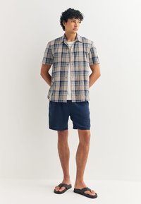 Short-sleeve plaid shirt in beige, blue, and green, paired with navy shorts and black flip-flops. Lightweight fabric with a relaxed fit.