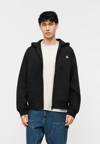 Νεαρός άνδρας που φοράει μαύρο φούτερ με φερμουάρ και εμπρόσθιες τσέπες πάνω από λευκό μπλουζάκι και μπλε τζιν, στέκεται μπροστά σε λευκό, απλό φόντο.