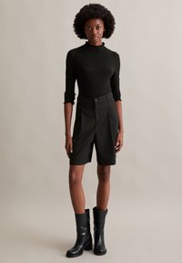 Schwarzes, geripptes Langarmoberteil mit Rüschenschnitt, kombiniert mit taillierten schwarzen Shorts und kniehohen schwarzen Stiefeln. Glatte Textur, figurbetontes Design.